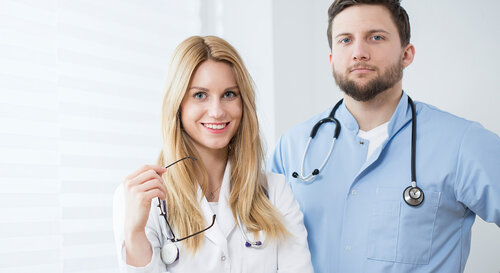 Eine junge Ärztin und ein junger Arzt stehen lächelnd in der Praxis nebeneinander